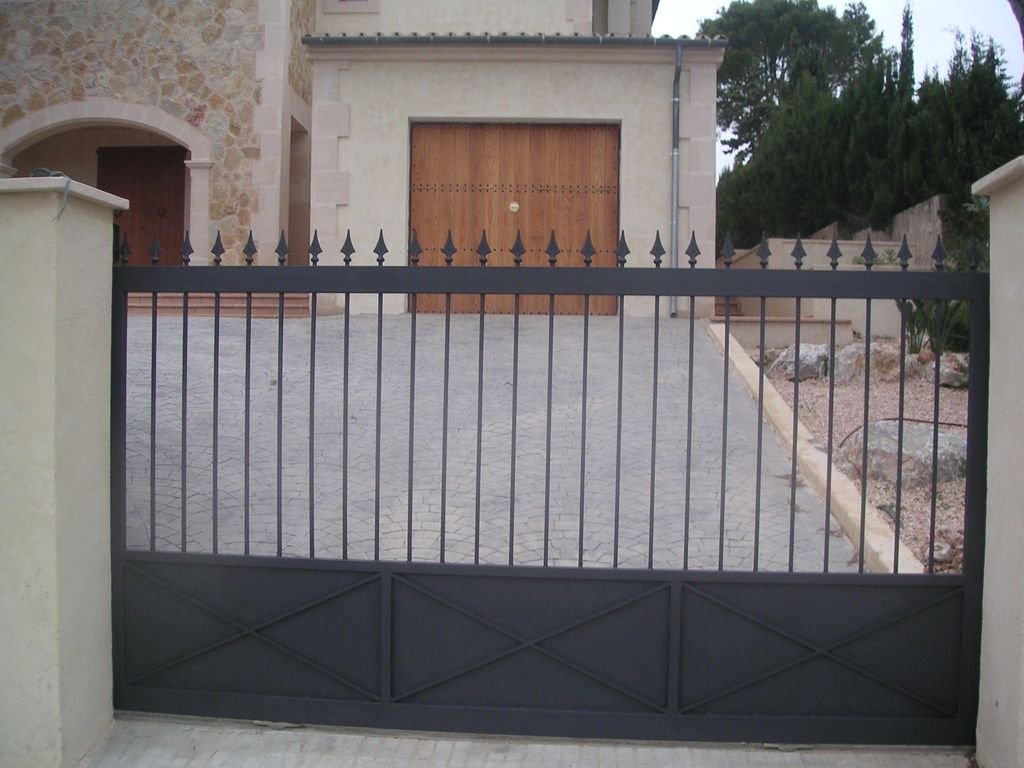 Portón negro con vallas en la entrada de una casa con cochera y jardín en el fondo