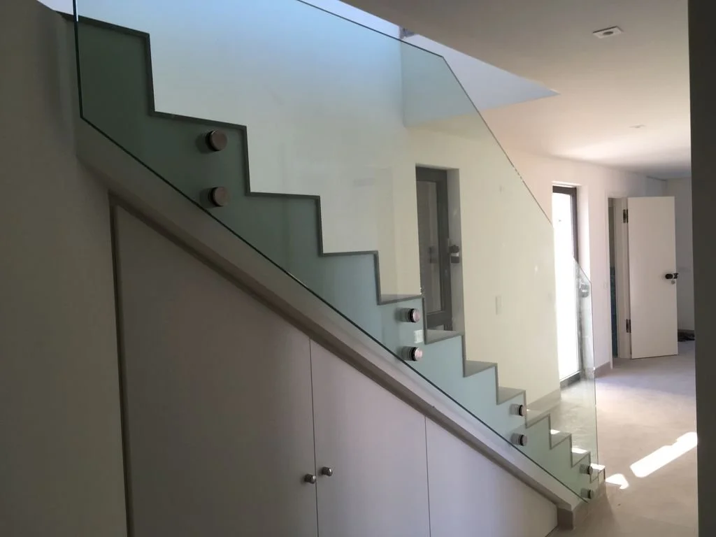 Escalera moderna con barandilla de vidrio y fijaciones negras en una habitación con puertas abiertas y luz solar entrando.