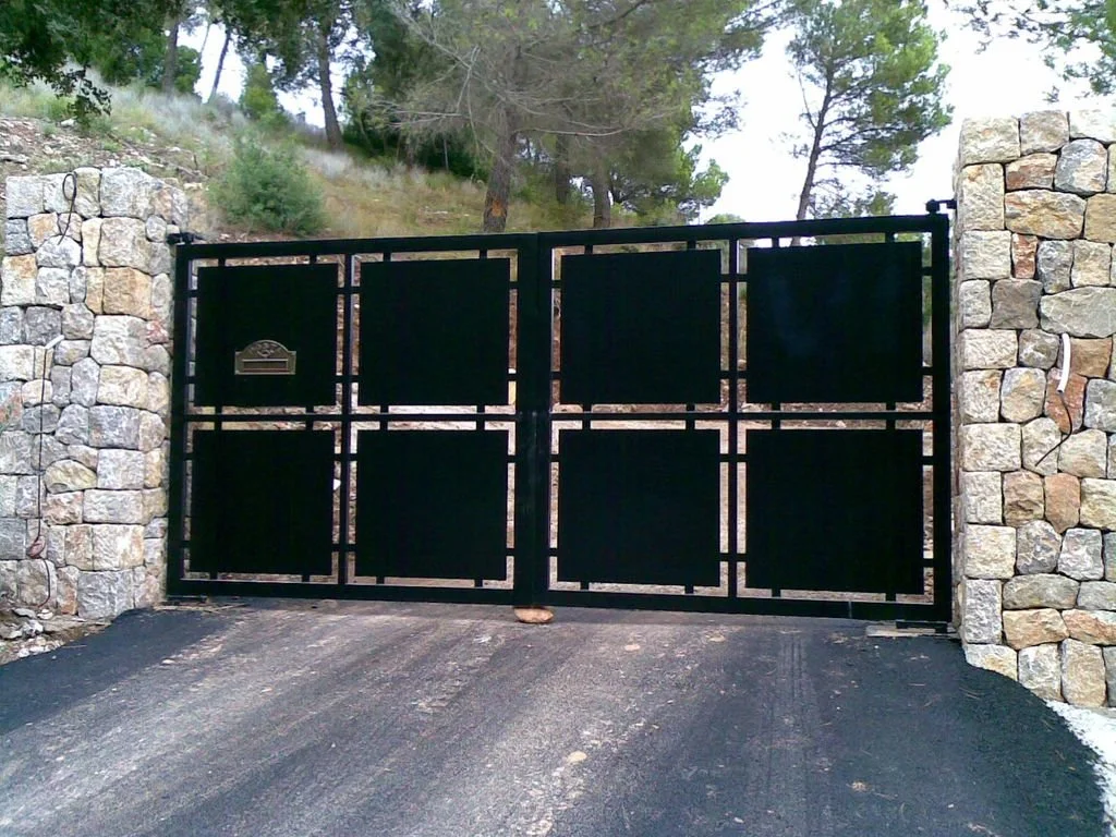 Puerta negra de puertas dobles con marco de línea recta y casillero, rodeada de muros de piedra, en una entrada de camino asfaltado, con árboles en el fondo.