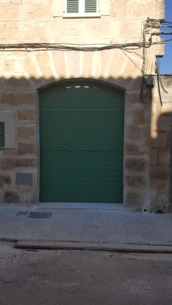 Puerta de garaje verde en fachada de piedra, ventanas con celosía y cables eléctricos en la parte superior.