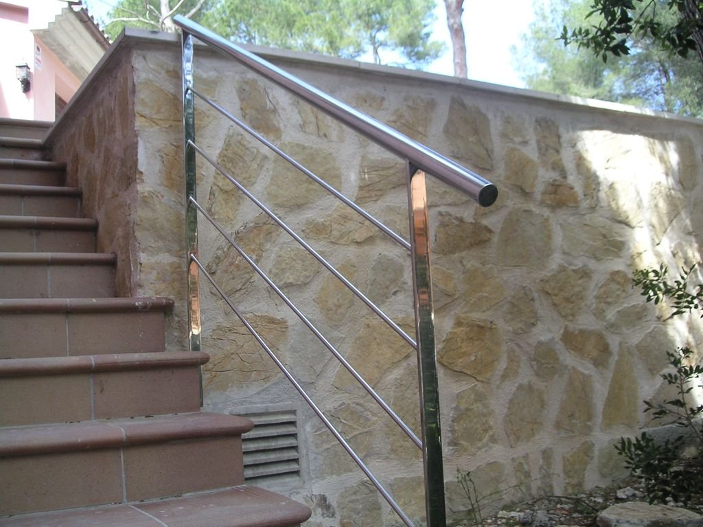 Escaleras de cerámica y una barandilla de metal junto a una pared de piedra, en un entorno exterior con árboles y cielo visible.