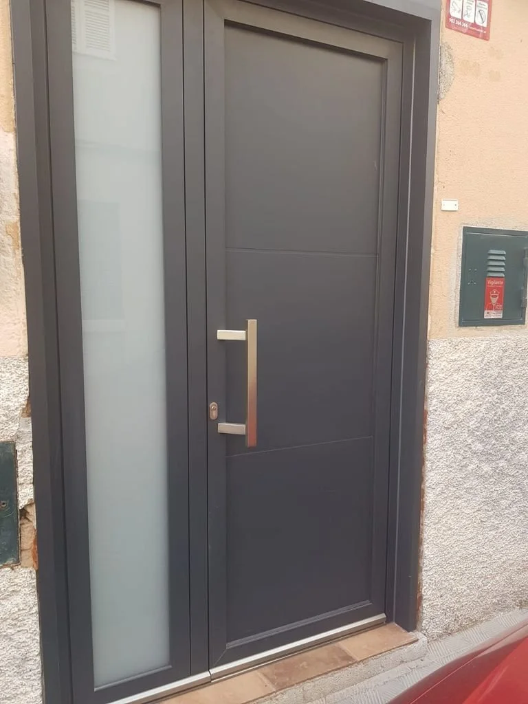 Puerta de entrada moderna de color gris oscuro con panel de cristal opaco a la izquierda, instalada en muro de piedra y yeso, con manija metálica larga a la izquierda.
