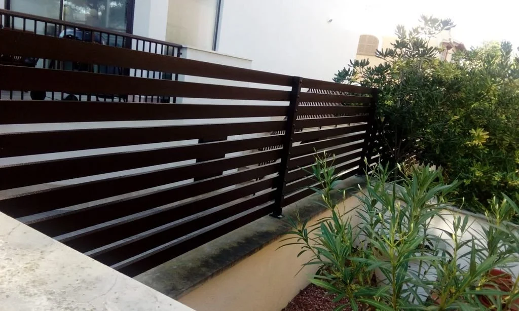 Vista de una cerca de madera oscura en un balcón con plantas verdes a un lado y un edificio en el fondo.