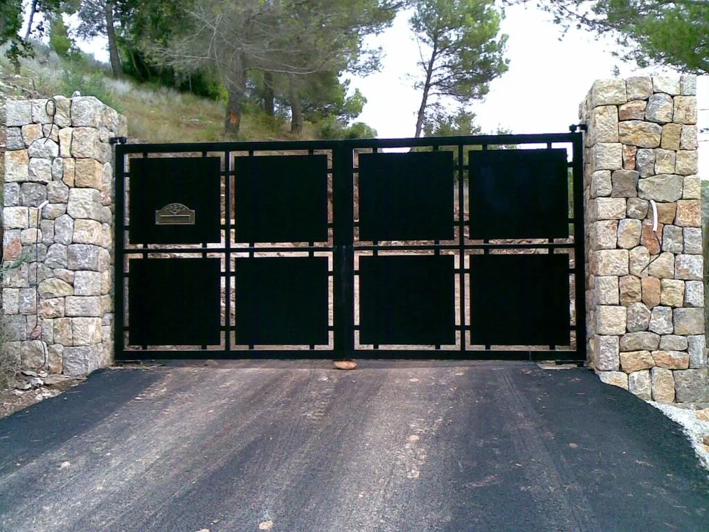 Puerta de garaje negra con marco de metal, flanqueada por pilares de piedra, en un entorno natural con árboles y césped.