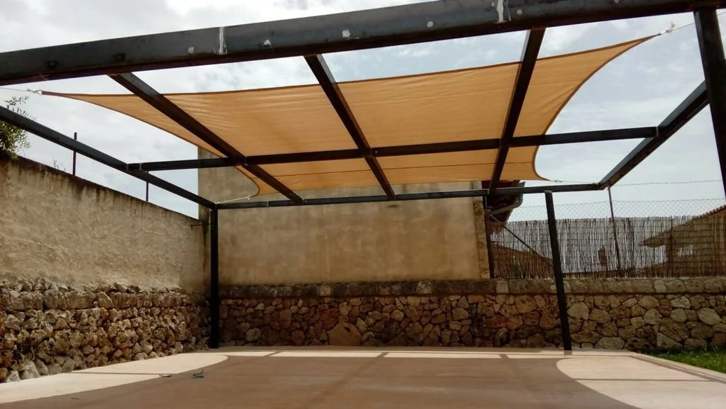Una estructura de acero con una lona beige en el techo, en un patio con paredes de piedra y muro de concreto, y un cielo nublado.
