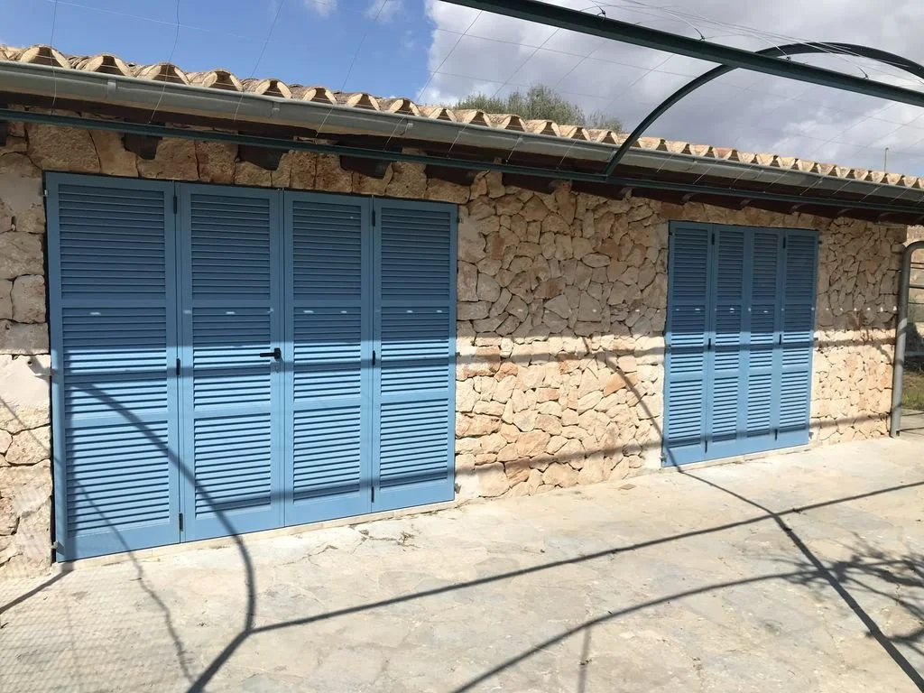 Casa de piedra con puertas y ventanas de persianas azules en un exterior con cielo parcialmente nublado.
