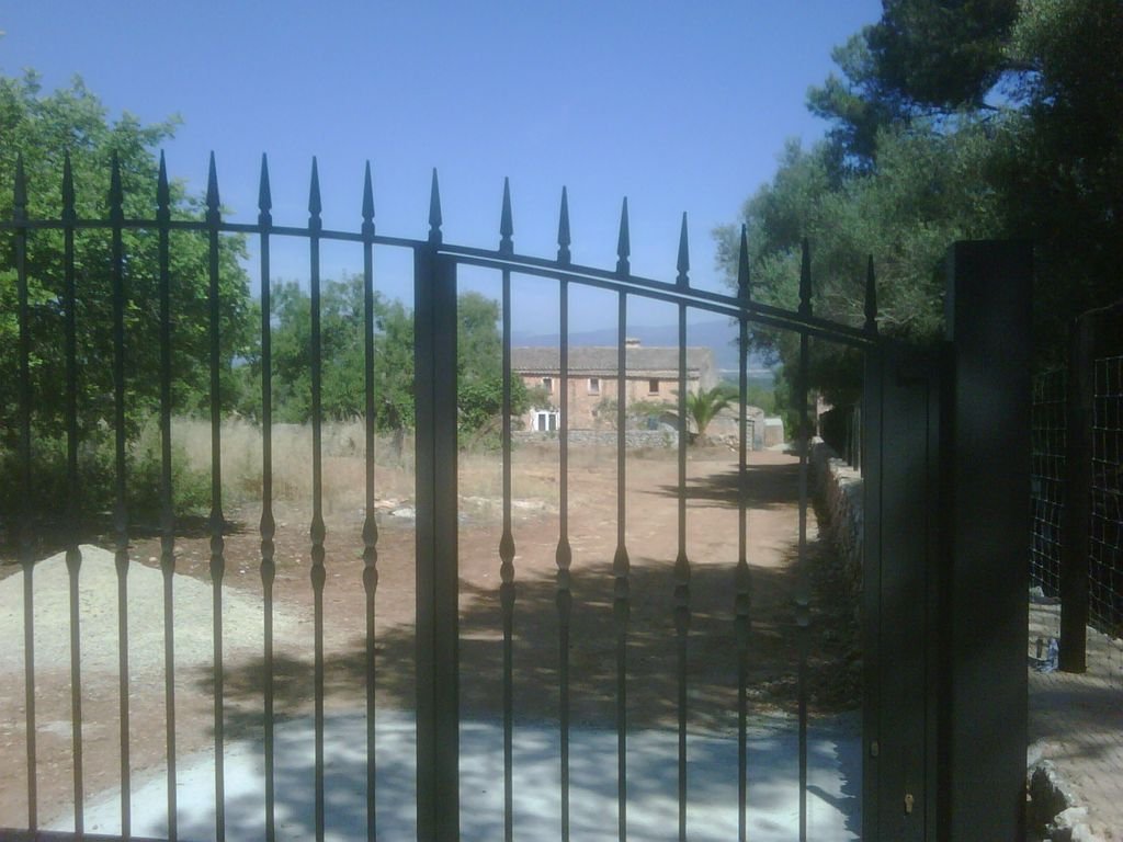 Puerta de herradura de metal negra que da a un camino con árboles y planta en un entorno rural, con un edificio en el fondo.