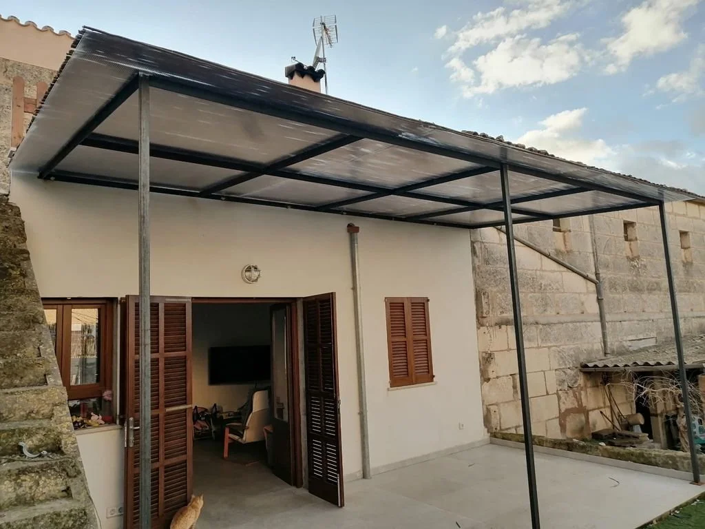 Patio exterior de una casa con una estructura metálica para techo, ventanas con persianas de madera y una puerta doble de madera que conduce a una sala internamente visible.