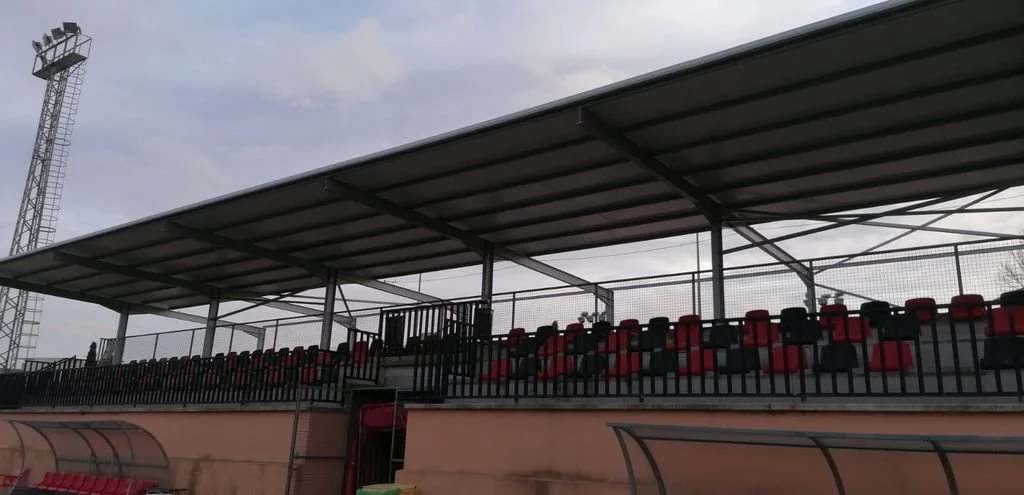Tribuna de estadio con asientos rojos y negros, techada y cercada con malla, en un día nublado.