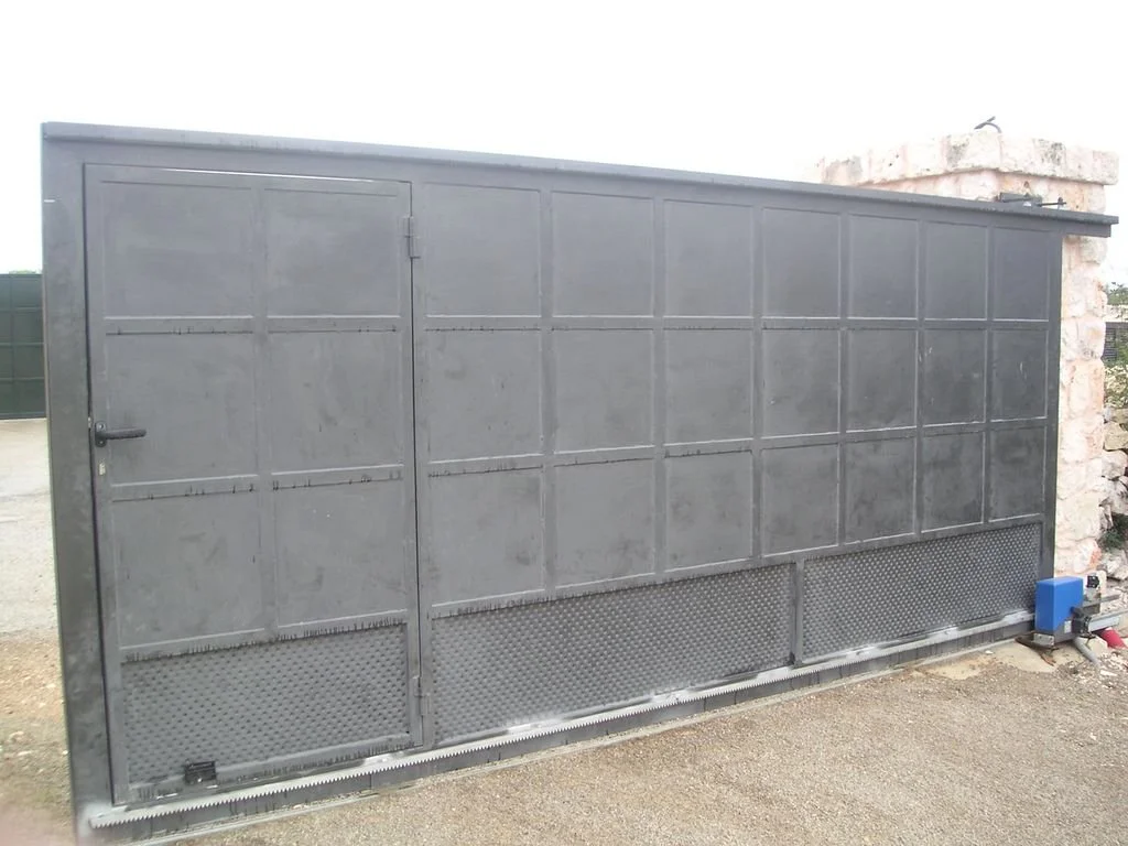 Puerta grande de metal gris con una pequeña puerta adicional en el lado izquierdo, ubicada en un espacio exterior.
