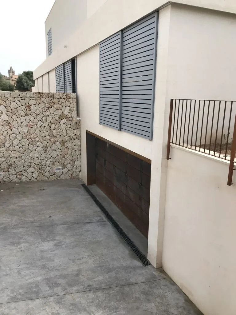Vista de una pared de edificio con ventanas con persianas grises y un muro de piedra en una esquina, con un suelo de cemento y una rejilla de drenaje en el suelo.