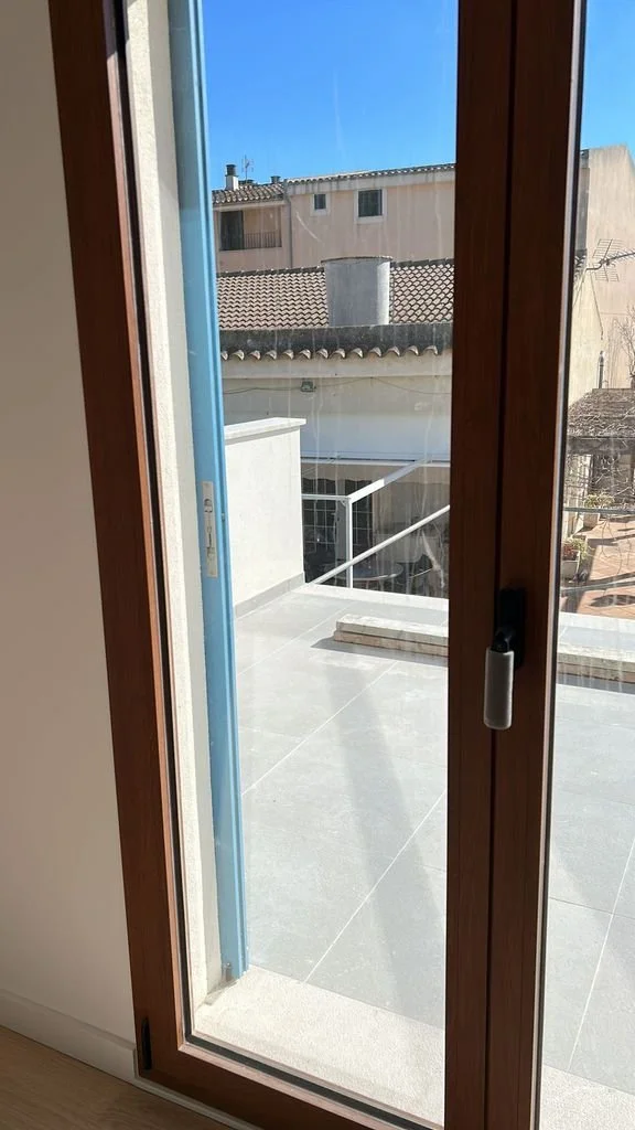Puerta de vidrio que da a una terraza con suelo de baldosas y barandilla de metal, con casas y cielo azul al fondo.