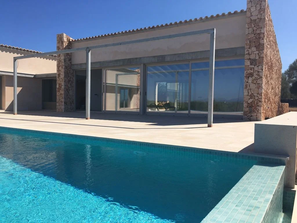 Casa moderna con fachada de piedra y amplia ventana de vidrio, alberca en primer plano, cielo despejado y área exterior de spa o terraza.