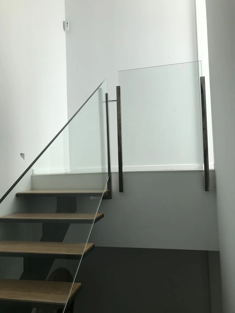 Escalera moderna de madera con barandal de vidrio y pasamanos metálicos en un interior minimalista.