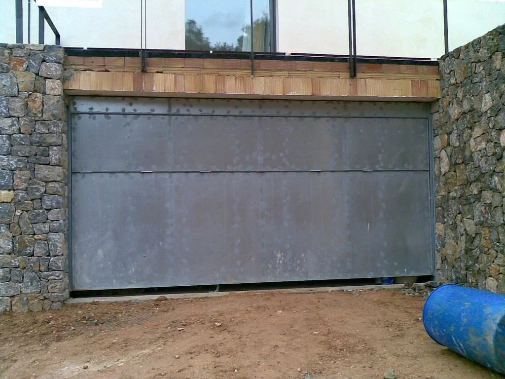 Puerta de garaje metálica enmarcada por muros de piedra y suelo de tierra, con una estructura de madera y vidrio en la parte superior.