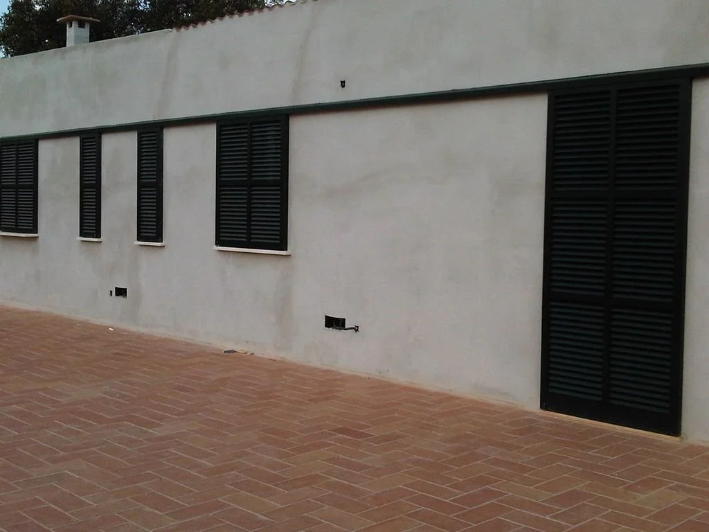 Pared de un edificio con cinco ventanas y una puerta, todas con persianas negras y rejas, y un piso de ladrillos.