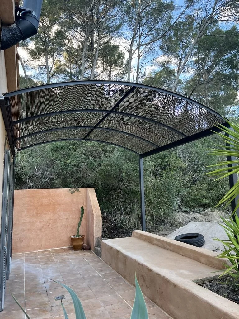 Un espacio exterior con techo de mimbre, una pared beige, plantas en macetas, una banca de concreto y un fondo con árboles.