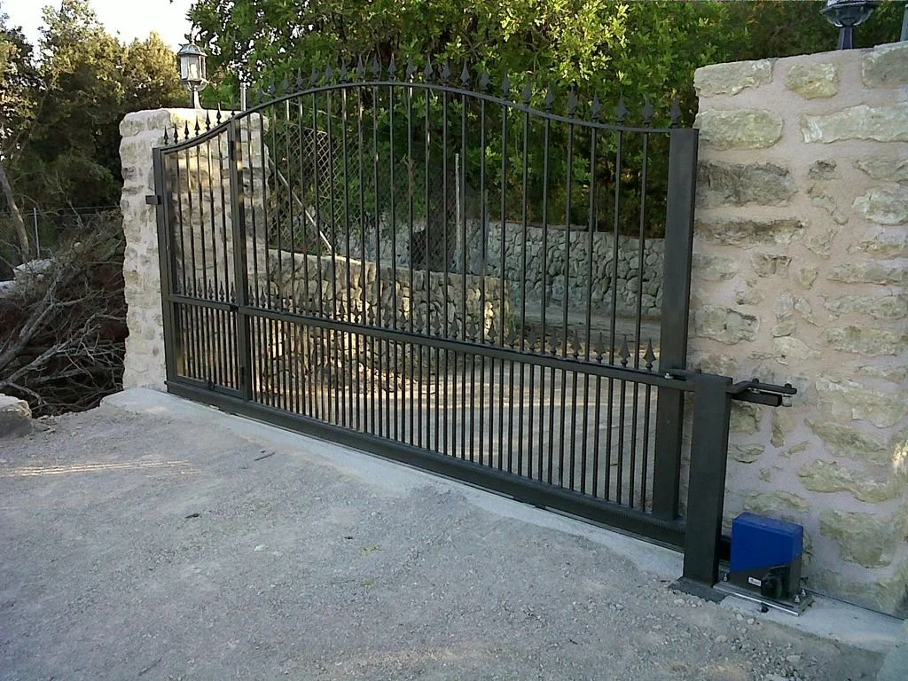 Puerta de rejas metálicas negra instalada entre dos muros de piedra, con un motor para apertura automática y un poste con luz en uno de los muros.