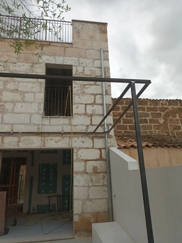 Construcción en proceso con estructura metálica y paredes de piedra, sin ventanas en el primer piso, y un cielo nublado.