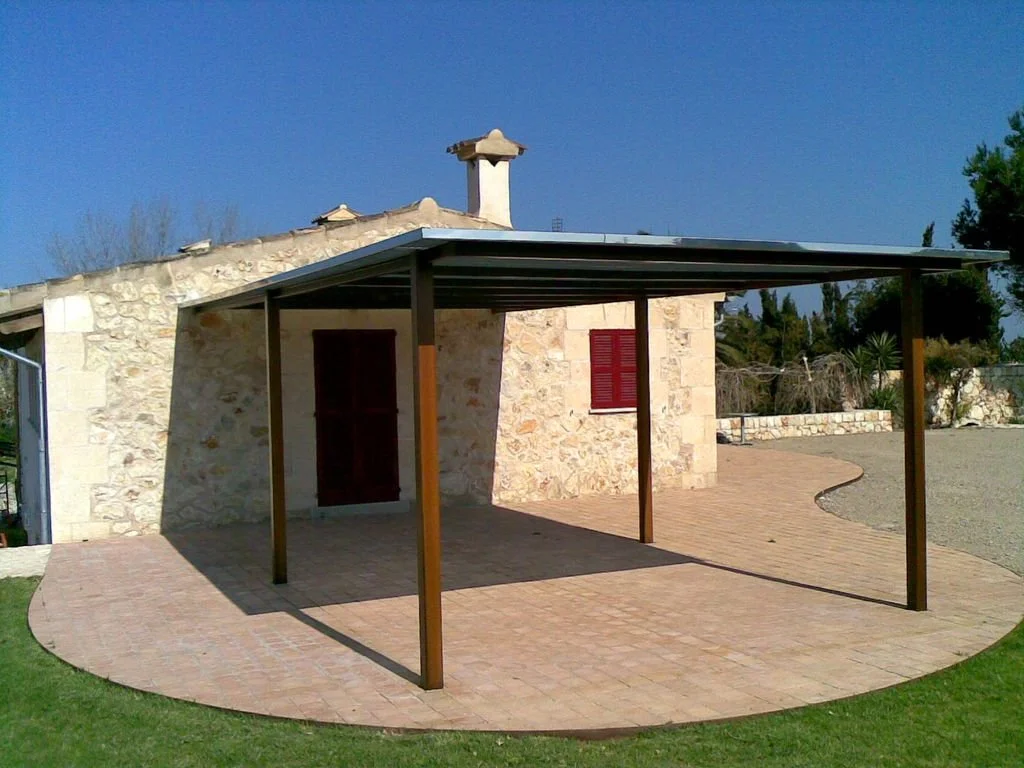 Casa de piedra con tejado de tejas y chimenea, con un toldo de madera y metal en el patio, rodeada de césped y camino de piedra.