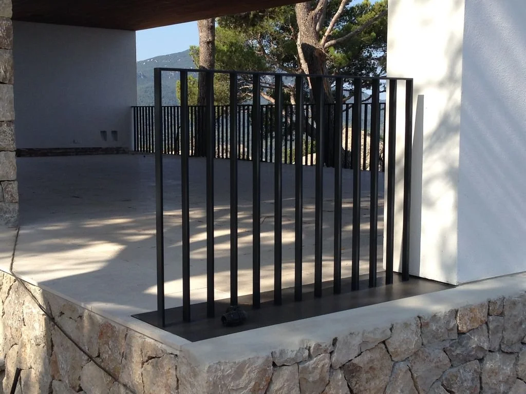 Baranda negra de metal en una terraza con muro de piedra y vista a árboles y montañas.