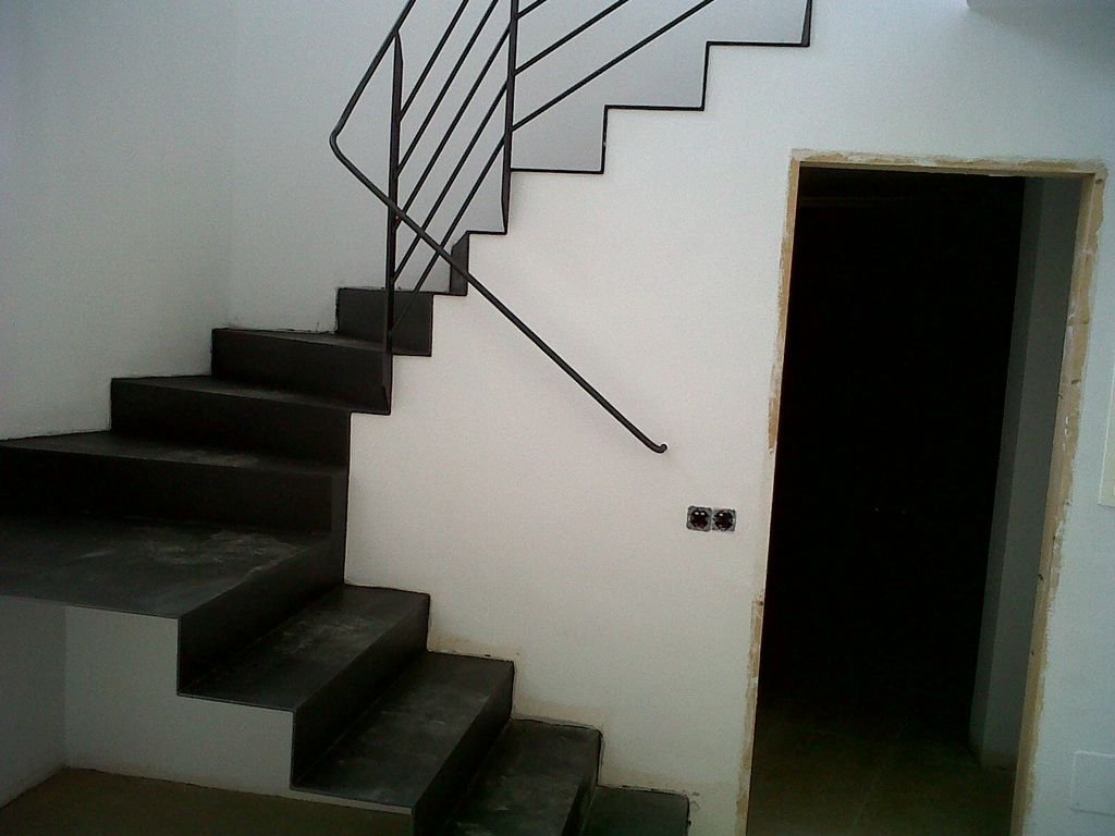 Escalera en construcción con barandilla de metal y paredes blancas, junto a una puerta sin marco en una habitación en obra.