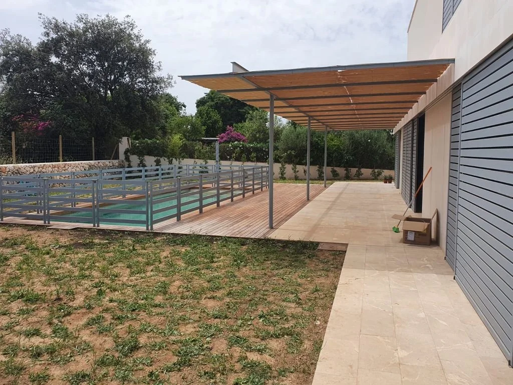 Área exterior de una casa con pasillo cubierto y suelo de baldosas, cerca de un espacio de tierra con plantas y árboles, con puertas enrollables en la fachada.