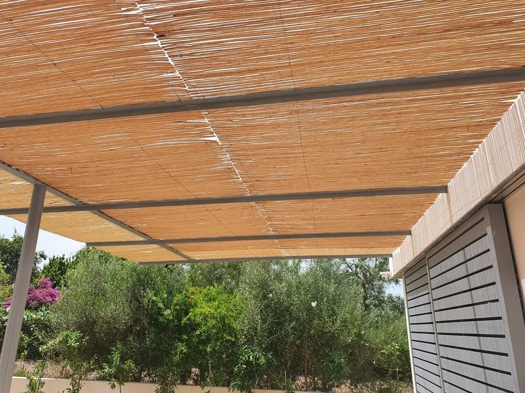 Persiana de bambú en un patio, con plantas y árboles verdes en el fondo.