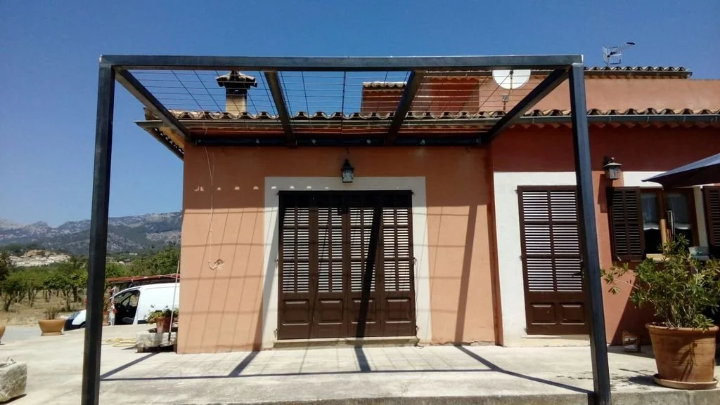 Casa de color terracota con puertas y ventanas de madera, estructura de metal en el frente, en un entorno rural con montañas en el fondo.