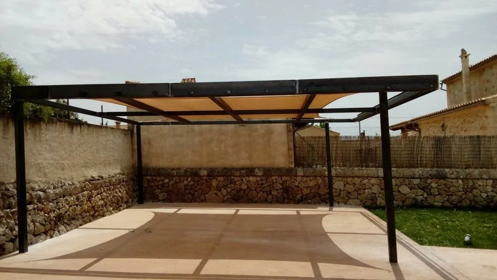 Estructura de pérgola en un patio, con sombra sobre el suelo de concreto y paredes de piedra y muro de ladrillos en el fondo.