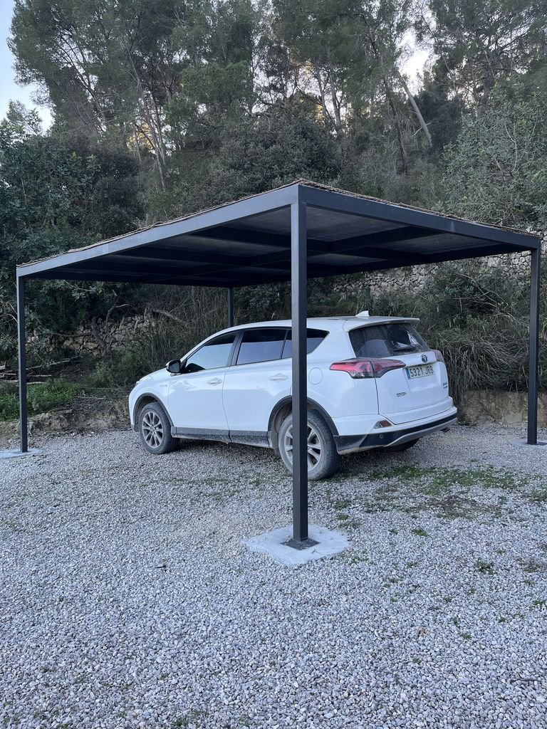 Cochera con estructura metálica y automóvil estacionado en un área con grava, rodeada de vegetación y árboles.