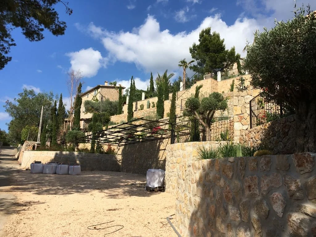 Vista de una casa en una loma con árboles y plantas, con muretes de piedra y cielo con nubes.