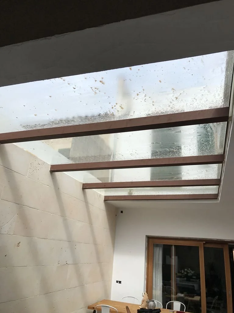 Interior de una casa con una claraboya en el techo, acero y una pared de ladrillos revestidos. Se puede ver una mesa de madera con sillas y una ventana grande con cortinas.