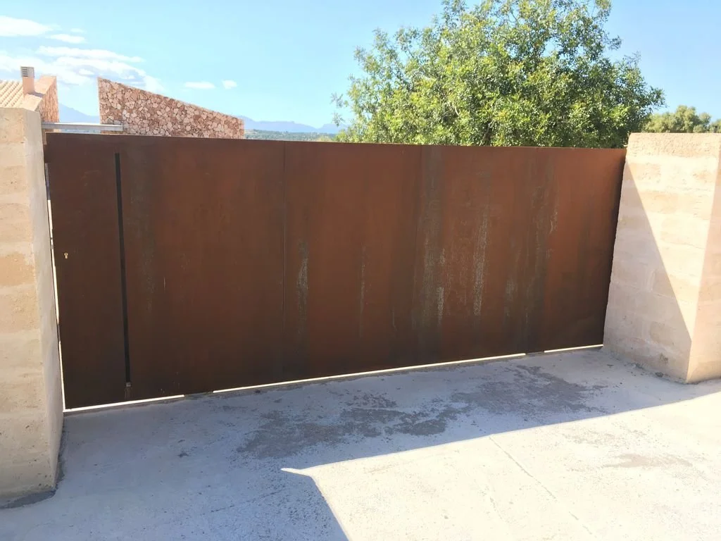 Puerta de metal oxidado en una pared de piedra, con árboles y cielo despejado en el fondo.
