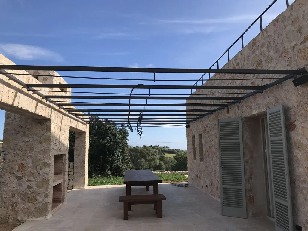 Estructura en construcción en una terraza de piedra con vista al campo, planchas de metal en el techo y ventanas con persianas verdes.