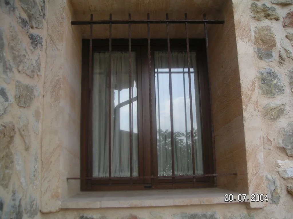 Ventana de madera con rejas de hierro en una pared de piedra, con cortinas blancas visibles desde afuera, fecha 20 de julio de 2004.