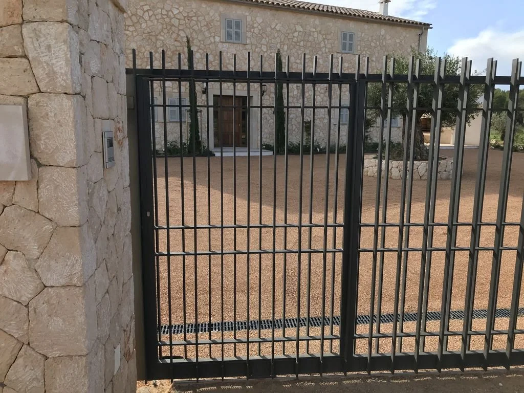 Puerta de reja negra en una entrada de casa de piedra, con jardín y árbol al fondo, en un día soleado.
