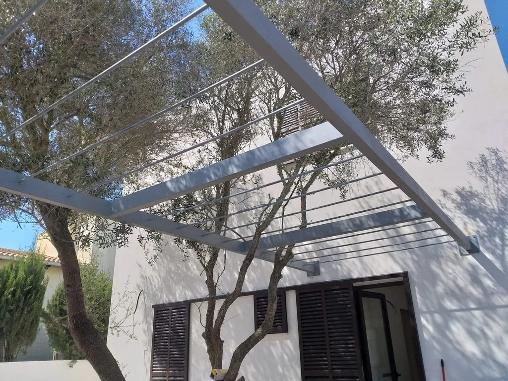Estructura metálica blanca con listones delgados en un patio junto a un árbol con ramas retorcidas, en frente de una pared blanca con ventanas con persianas oscuras.