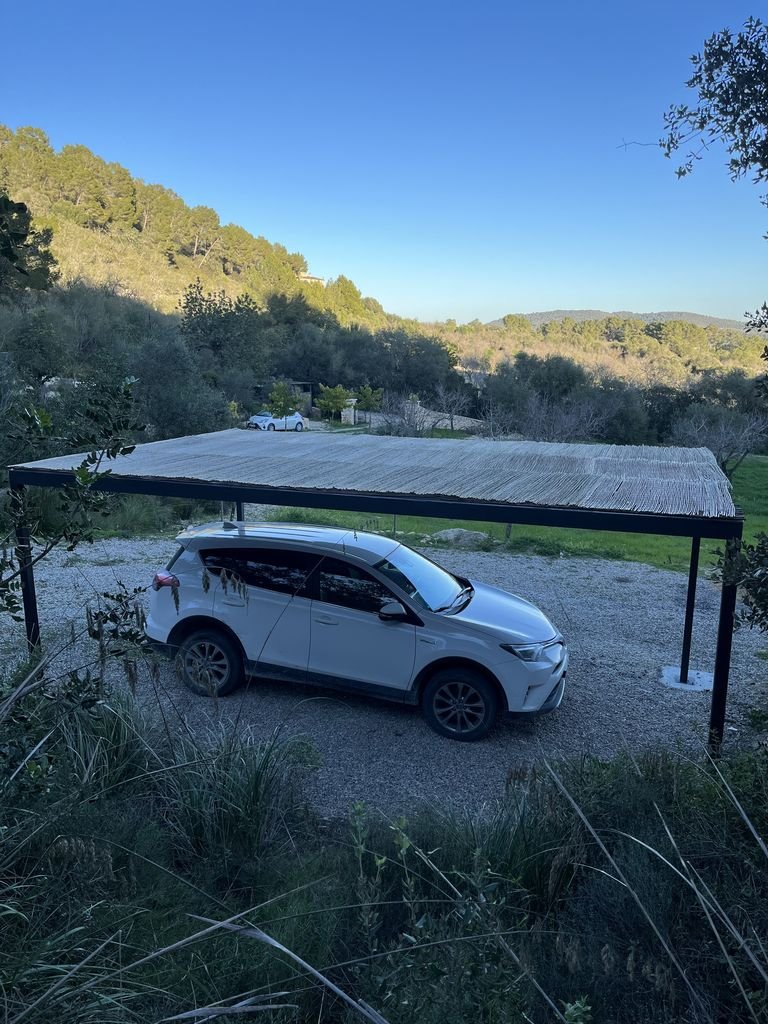 Cochera al aire libre con techo de lámina, en un entorno natural rodeado de árboles y colinas, con un auto estacionado.