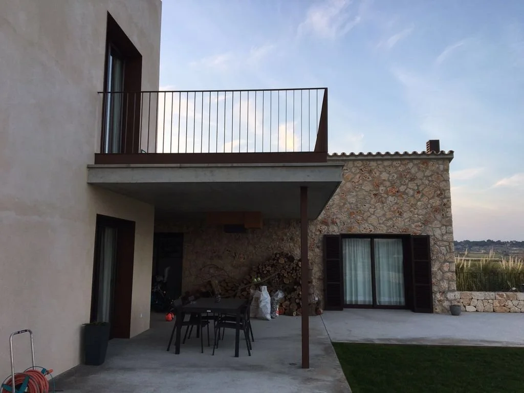 Vista de un patio de una casa moderna con balcón, ventanas con cortinas, pared de piedra, y un área de asador o leña con muebles de exterior.