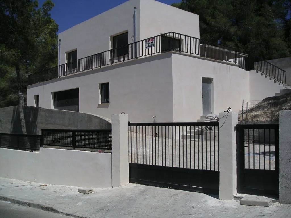 Casa moderna blanca con muro de entrada y puertas de rejas negras, en un entorno con árboles y terreno en pendiente.
