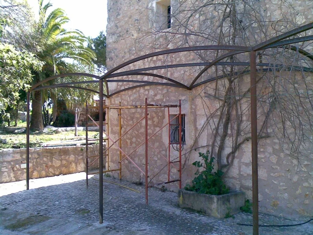 Estructura metálica en construcción frente a una pared de piedra, con andamios en el centro y vegetación en el fondo.