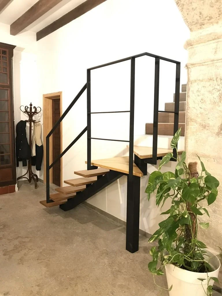 Escalera moderna con barandal negro, pasos de madera y una planta en una maceta blanca al lado, en el interior de una casa.