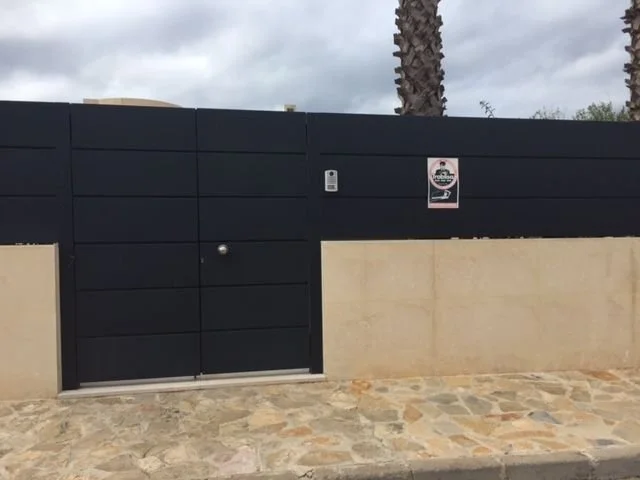 Puerta de metal negro con muro de bloques de concreto y pared de madera, cartel de advertencia y palma en el fondo, cielo nublado.
