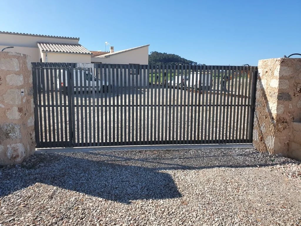 Puerta metálica negra con rejas en una entrada de piedra y grava, con casas y montañas al fondo, bajo cielo despejado.