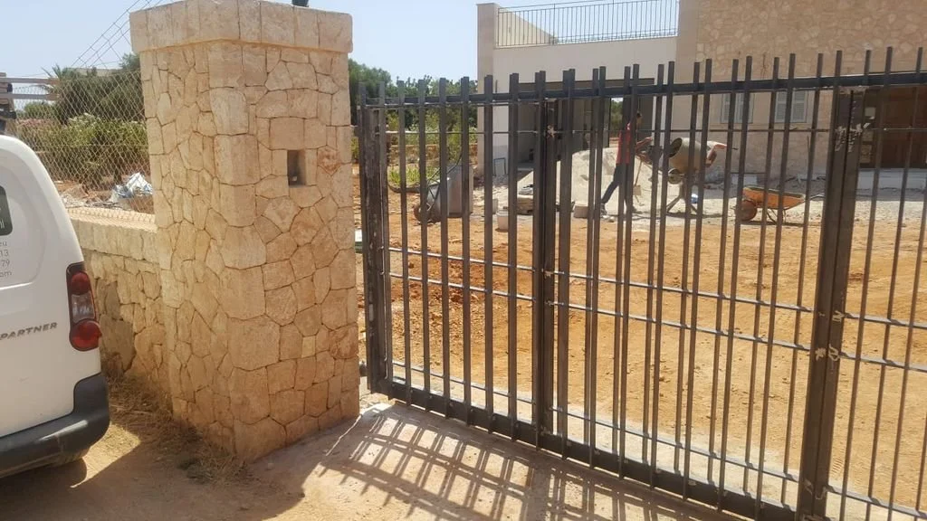 Puerta de reja negra en un muro de piedra que da a un terreno de tierra con construcción y personas en movimiento.
