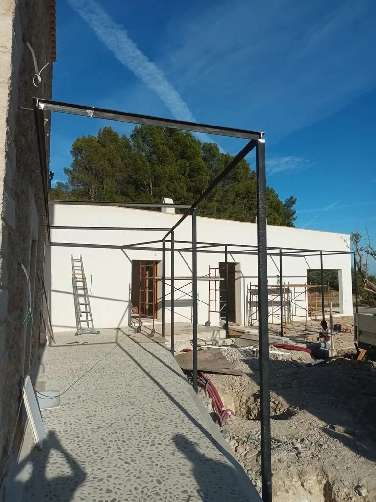 Construcción de una casa con estructura metálica y paredes blancas, escaleras y herramientas en el sitio, andamios y una persona trabajando en el fondo.
