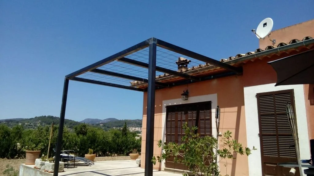 Casa de color rosa con ventanas de madera con persianas, techo de tejas, televisión satelital y mobiliario exterior en un patio con plantas y vista a las montañas