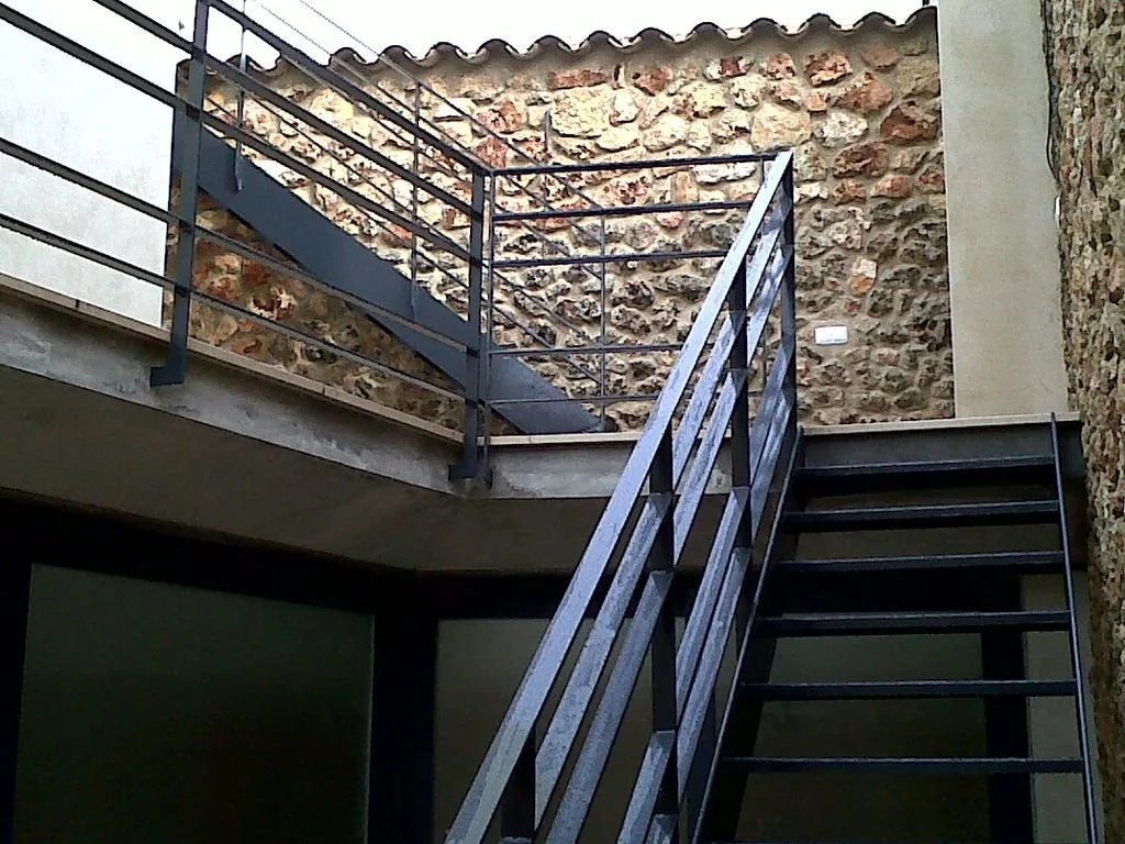 Escalera de metal en un patio con paredes de piedra y muro de ladrillos a la vista.