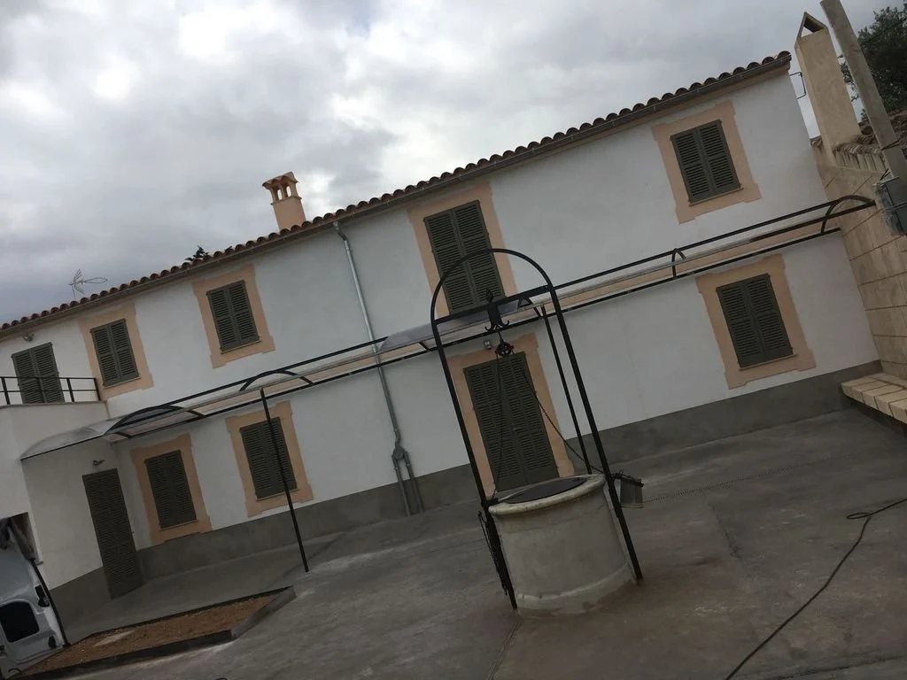 Edificio de dos pisos con ventanas con persianas verdes, en un patio con mástil de pozo antiguo en el centro, cielo nublado y paredes blancas con marcos de color beige.